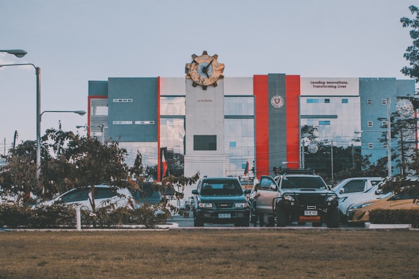 A modern building with a large emblem at the top is visible; it features gray, red, and blue tones. In front of the building, there is a row of parked cars and some greenery, with a lawn in the foreground. The building has a slogan about innovation visible on its facade.