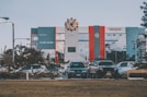 A modern building with a large emblem at the top is visible; it features gray, red, and blue tones. In front of the building, there is a row of parked cars and some greenery, with a lawn in the foreground. The building has a slogan about innovation visible on its facade.