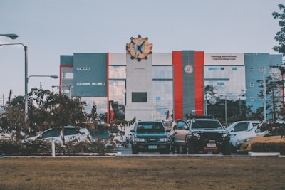 A modern building with a large emblem at the top is visible; it features gray, red, and blue tones. In front of the building, there is a row of parked cars and some greenery, with a lawn in the foreground. The building has a slogan about innovation visible on its facade.