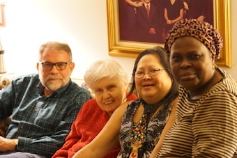 Four people are sitting closely together on a couch inside a well-lit room. They appear to be diverse in age and ethnicity, smiling warmly at the camera. Behind them, there is a large framed portrait on the wall.