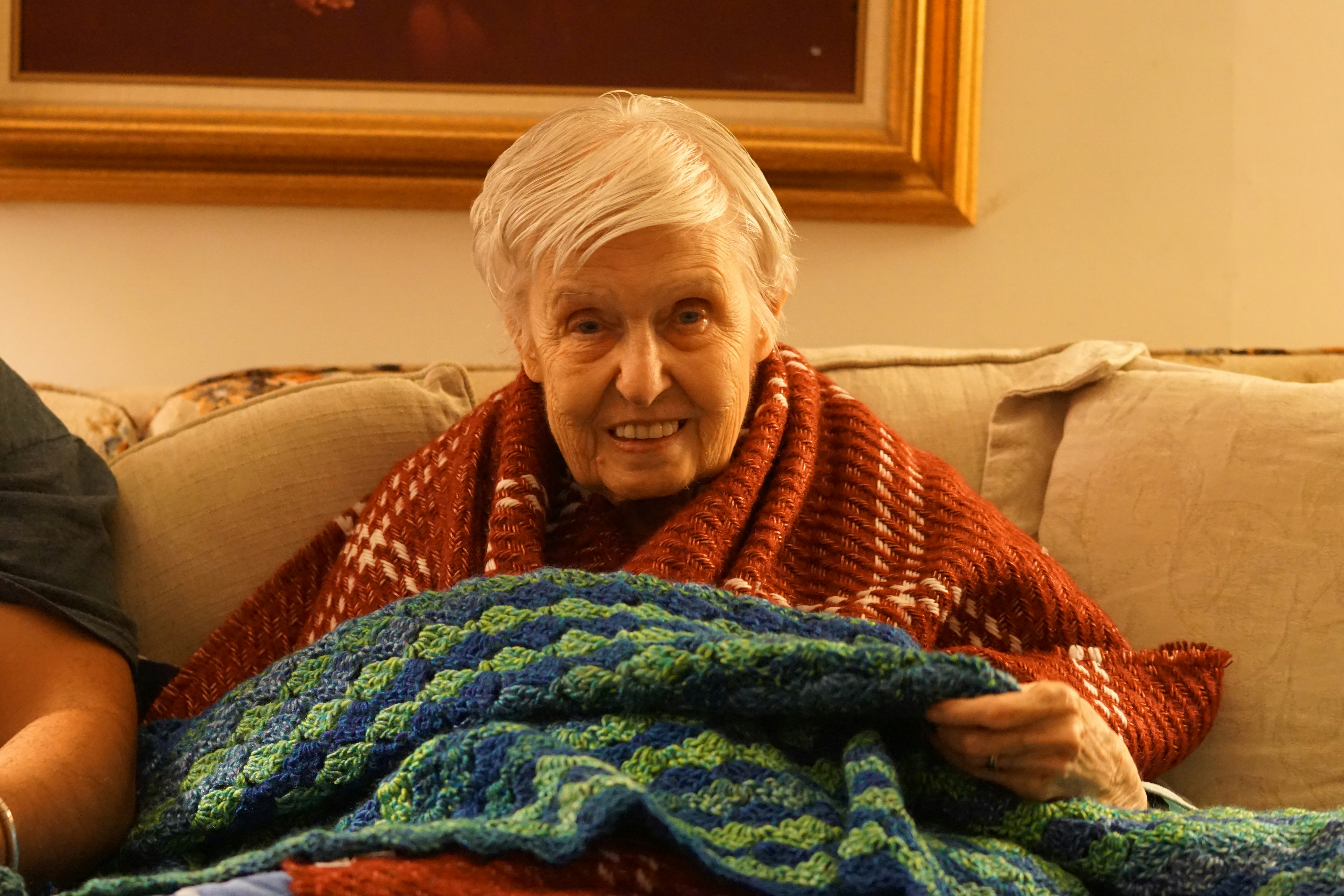Elderly woman sitting alone in a cold-looking living room with a blanket