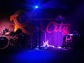 A dimly lit stage with musical instruments, including a drum set, two electric guitars, and a keyboard. The backdrop features the words 'Rock City' in bold letters with a stylized design. The lighting creates a moody ambiance, with blue and purple hues dominating the scene.