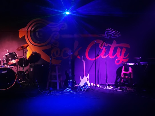 A dimly lit stage with musical instruments, including a drum set, two electric guitars, and a keyboard. The backdrop features the words 'Rock City' in bold letters with a stylized design. The lighting creates a moody ambiance, with blue and purple hues dominating the scene.