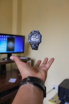 A digital watch floats above an outstretched hand in an indoor setting. A computer monitor is visible in the background on a wooden desk, and a piece of paper is on the surface. The setting appears to be an office or study room with beige walls.
