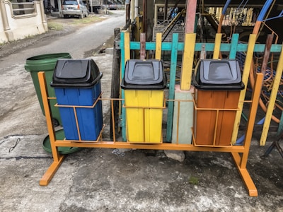 Three trash bins in blue, yellow, and orange are mounted on a metal frame. They are located on a concrete surface with colorful wooden fencing in the background.