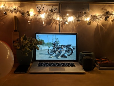 A cozy desk setup with warm lighting is decorated with a string of lights and stars. A laptop screen displays a photograph of a person sitting next to a motorcycle. Surrounding the computer are various items including a green mug, two books, a potted plant, a smartphone, and a cricket bat.