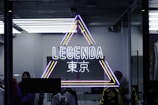 A neon sign is displayed in a shop window, featuring a triangle outlined with bright yellow, pink, and blue colors. Inside the triangle, the word 'LEGENDA' is illuminated in white neon lights above some Japanese characters. Within the store, clothing items are visible alongside a mannequin. The environment inside the shop appears modern and stylish, with a hint of industrial design from the visible ceiling and lighting fixtures.