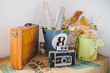 A vintage camera is placed on a burlap sack alongside a wooden suitcase. Next to them, a metal bucket with a painted number eight holds rolled-up papers and yellow dried flowers. A green watering can with a distressed appearance sits to the right, adding to the rustic aesthetic.