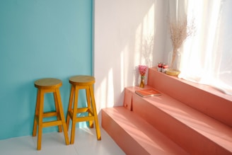 A room corner features two tall wooden stools with round seats placed against a soft teal wall. Adjacent to them are steps painted in a muted coral color, under soft, diffused natural light from a nearby window with sheer white curtains. Atop the steps are decorative items including a vase filled with pink flowers, a couple of stacked books with red covers, and a few canned goods. Another taller vase with dried branches is positioned against the wall, creating an inviting and serene atmosphere.