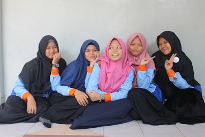 Five young people are sitting on the floor, leaning against a light-colored wall. They are wearing uniforms consisting of blue shirts with orange cuffs, and hijabs in various colors including black, blue, and pink. They appear to be in a relaxed and cheerful mood, with some making peace signs with their hands.