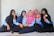 Five young people are sitting on the floor, leaning against a light-colored wall. They are wearing uniforms consisting of blue shirts with orange cuffs, and hijabs in various colors including black, blue, and pink. They appear to be in a relaxed and cheerful mood, with some making peace signs with their hands.