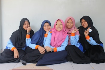 Five young people are sitting on the floor, leaning against a light-colored wall. They are wearing uniforms consisting of blue shirts with orange cuffs, and hijabs in various colors including black, blue, and pink. They appear to be in a relaxed and cheerful mood, with some making peace signs with their hands.