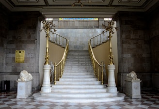 A grand, elegant staircase with white marble steps and ornate golden railings, flanked by two stone lion statues. The staircase is set in a classic interior with checkered flooring and stone walls, leading up to a well-lit area above. The intricate ceiling features plaster designs and a stained glass panel can be seen at the top.