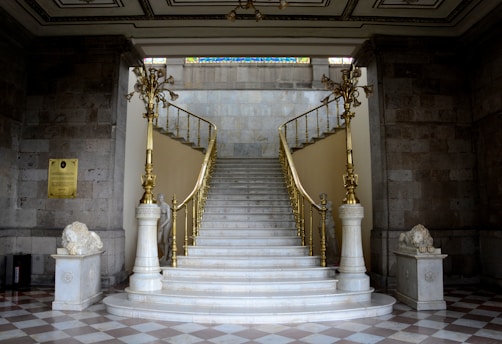 A grand, elegant staircase with white marble steps and ornate golden railings, flanked by two stone lion statues. The staircase is set in a classic interior with checkered flooring and stone walls, leading up to a well-lit area above. The intricate ceiling features plaster designs and a stained glass panel can be seen at the top.