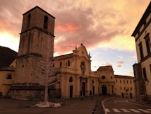 A historic church building with an imposing stone tower is set against a vibrant sunset sky, creating a striking contrast between the warm tones of the architecture and the vivid colors of the sky. The foreground features a small courtyard with a barren tree, and the surrounding buildings exhibit classic architectural features.