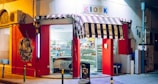 A small, brightly lit kiosk with a striped awning is situated on a quiet street corner. The storefront is painted red with artistic graffiti on the wall. Shelves inside are filled with various snacks and products. A large sign reading 'KIOSK' is displayed above the entrance.