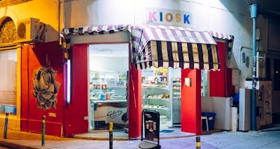 A small, brightly lit kiosk with a striped awning is situated on a quiet street corner. The storefront is painted red with artistic graffiti on the wall. Shelves inside are filled with various snacks and products. A large sign reading 'KIOSK' is displayed above the entrance.