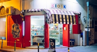 A small, brightly lit kiosk with a striped awning is situated on a quiet street corner. The storefront is painted red with artistic graffiti on the wall. Shelves inside are filled with various snacks and products. A large sign reading 'KIOSK' is displayed above the entrance.