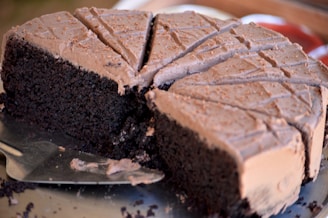 A richly textured chocolate cake with a smooth, creamy frosting on top. Several slices have been cut, revealing the dark and moist interior of the cake. The cake appears to be freshly baked, with a serving utensil nearby.