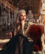 A woman with long blonde hair sits elegantly on a plush red chair wearing a golden crown and shimmering gold and black dress. The background features an opulent hall with large mirrors, chandeliers, and intricate gold details, suggesting a palace setting.