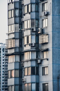 A close-up view of a modern residential building with gray tiled exterior walls. Multiple windows display a variety of curtains and reflect soft ambient light. Several air conditioning units are mounted outside, creating a pattern on the façade.