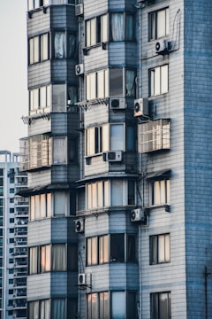 A close-up view of a modern residential building with gray tiled exterior walls. Multiple windows display a variety of curtains and reflect soft ambient light. Several air conditioning units are mounted outside, creating a pattern on the façade.