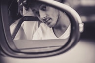 A young man with a thoughtful expression is reflected in a car's side mirror, captured in black and white. He is wearing a light-colored shirt, and his head tilts slightly as he looks towards the camera.