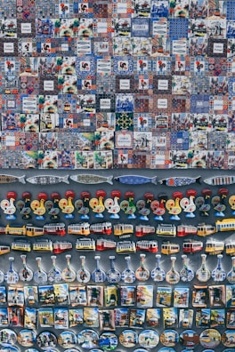 A collection of colorful and varied souvenirs arranged on display. The top section features a series of tiles with intricate patterns and illustrations, many labeled with 'Lisboa' or 'Lisbon Portugal'. Below the tiles, there are magnets shaped like roosters, trams, and traditional bottles with detailed designs. The display exudes a sense of Portuguese culture and heritage.