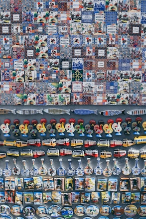 A collection of colorful and varied souvenirs arranged on display. The top section features a series of tiles with intricate patterns and illustrations, many labeled with 'Lisboa' or 'Lisbon Portugal'. Below the tiles, there are magnets shaped like roosters, trams, and traditional bottles with detailed designs. The display exudes a sense of Portuguese culture and heritage.