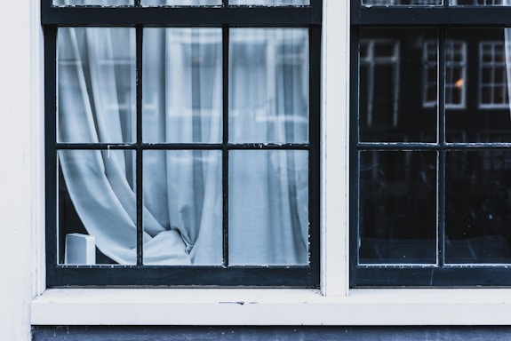A window with multiple panes has sheer white curtains partially drawn, creating a soft and tranquil atmosphere. The window frame is painted black with a slightly worn appearance. Light from outside casts soft shadows on the curtains.