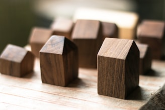 A collection of small, wooden blocks with simple geometric shapes and smooth surfaces is arranged on a light wooden surface. The blocks vary in size and are positioned in a way that some of them appear slightly out of focus, creating a sense of depth.