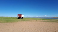 A red truck with a white cargo area is parked on an open plain with sparse grass and dirt. A clear blue sky extends above, with mountains visible in the background near a lake. The landscape is vast and uninhabited, conveying a sense of solitude and wilderness.