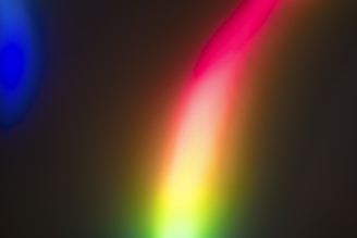 A vibrant and abstract display of colors blending together, predominantly featuring a bright spectrum of red, pink, yellow, and green. The background is dark, creating a stark contrast with the luminous, rainbow-like streaks of light.