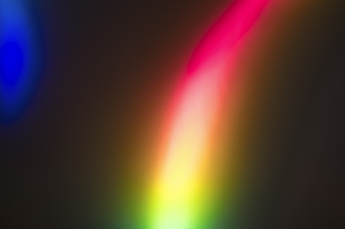 A vibrant and abstract display of colors blending together, predominantly featuring a bright spectrum of red, pink, yellow, and green. The background is dark, creating a stark contrast with the luminous, rainbow-like streaks of light.