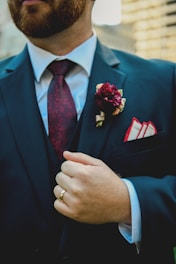 A person wearing a dark suit with a white shirt and a patterned maroon tie. A maroon flower boutonniere is pinned on the left lapel above a pocket square. The person is also wearing a gold ring and is seen holding the suit lapel.
