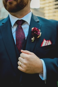 A person wearing a dark suit with a white shirt and a patterned maroon tie. A maroon flower boutonniere is pinned on the left lapel above a pocket square. The person is also wearing a gold ring and is seen holding the suit lapel.
