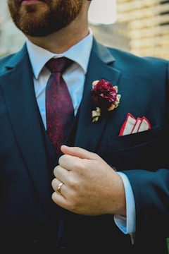 A person wearing a dark suit with a white shirt and a patterned maroon tie. A maroon flower boutonniere is pinned on the left lapel above a pocket square. The person is also wearing a gold ring and is seen holding the suit lapel.