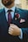 A person wearing a dark suit with a white shirt and a patterned maroon tie. A maroon flower boutonniere is pinned on the left lapel above a pocket square. The person is also wearing a gold ring and is seen holding the suit lapel.