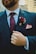 A person wearing a dark suit with a white shirt and a patterned maroon tie. A maroon flower boutonniere is pinned on the left lapel above a pocket square. The person is also wearing a gold ring and is seen holding the suit lapel.
