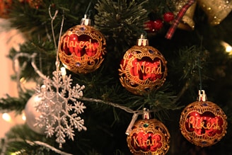 Ornate Christmas tree with personalized red and gold ornaments featuring names, surrounded by green pine branches and decorated with a silver snowflake and string lights.
