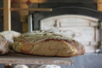 Freshly baked bread with a golden crust resting on a wooden paddle in front of a rustic oven. The bread appears soft and fluffy, indicative of a perfect bake. The background is slightly blurred, focusing the attention on the bread and the warm ambiance of a traditional bakery.