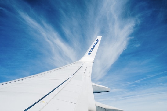 A Ryanair airplane wing seen from a passenger's perspective, set against a backdrop of a clear blue sky with wispy clouds. The wing extends diagonally across the image, emphasizing the vastness of the sky.
