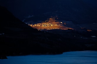 A small settlement is illuminated by a patch of sunlight amidst a vast, dark landscape. The settlement is situated on a hillside with a clear view of a body of water in the foreground. Most of the surrounding landscape, including mountains and additional structures, is in shadow, creating a stark contrast with the brightly lit area.