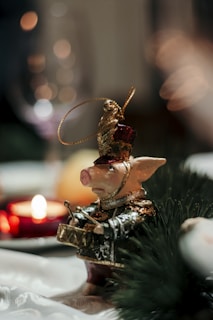 A decorative figurine of a pig dressed as a soldier stands on a table. It has a drum, a small hat adorned with a golden plume, and wears a detailed military uniform with gold accents. In the background, there are bokeh lights and a red candle adding a warm glow to the scene.