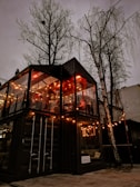 A cozy two-story structure made from shipping containers, adorned with warm string lights, sits beneath the bare branches of tall trees. The interior glows with ambient illumination, suggesting a welcoming atmosphere inside.