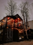 A cozy two-story structure made from shipping containers, adorned with warm string lights, sits beneath the bare branches of tall trees. The interior glows with ambient illumination, suggesting a welcoming atmosphere inside.