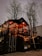 A cozy two-story structure made from shipping containers, adorned with warm string lights, sits beneath the bare branches of tall trees. The interior glows with ambient illumination, suggesting a welcoming atmosphere inside.