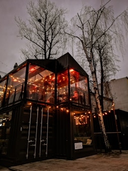 A cozy two-story structure made from shipping containers, adorned with warm string lights, sits beneath the bare branches of tall trees. The interior glows with ambient illumination, suggesting a welcoming atmosphere inside.