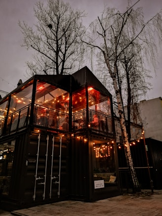 A cozy two-story structure made from shipping containers, adorned with warm string lights, sits beneath the bare branches of tall trees. The interior glows with ambient illumination, suggesting a welcoming atmosphere inside.
