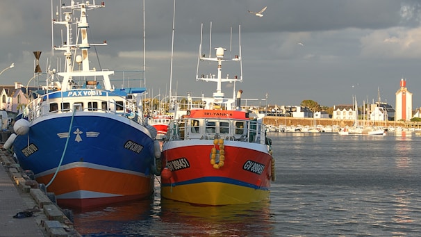 Two fishing boats docked at a harbor, one painted in bright blue and orange and the other in red and yellow, are positioned side by side. The harbor is lined with small buildings, and a lighthouse with red accents stands prominently. A few seagulls are flying in the cloudy sky above.